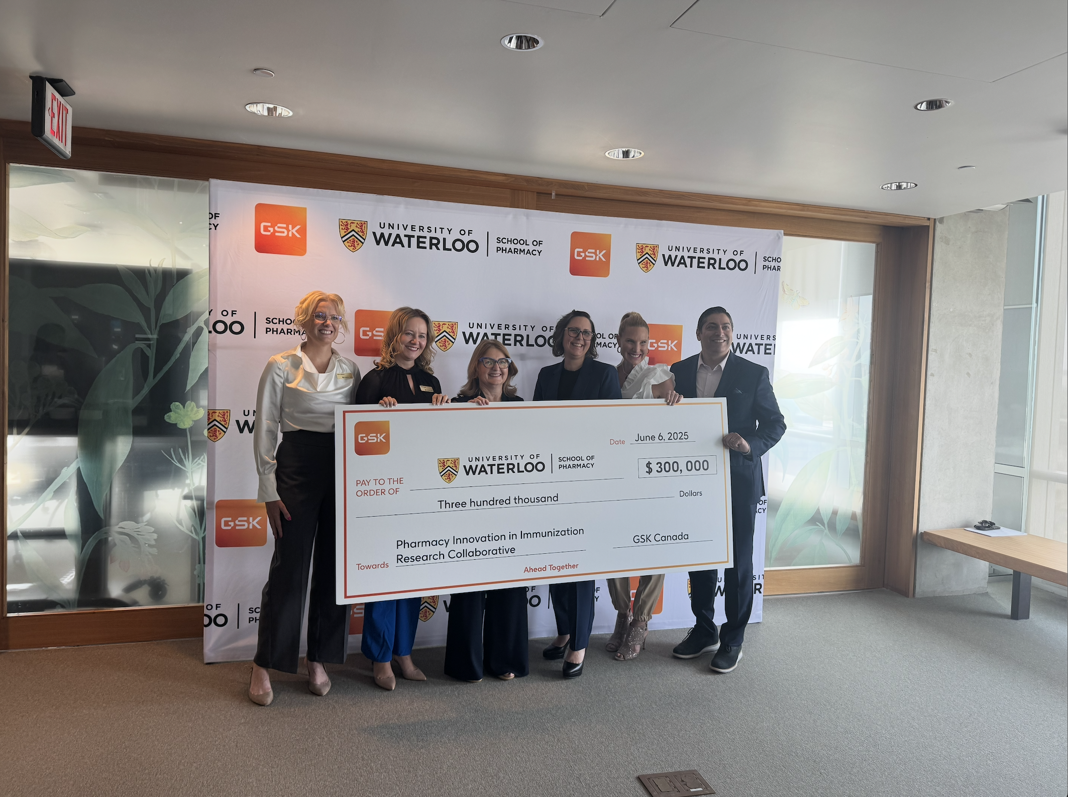 Five people stand smiling indoors, holding an oversized $300,000 check from GSK Canada, awarded to the School of Pharmacy at University of Waterloo for their immunization initiative. University and GSK logos are displayed behind them.